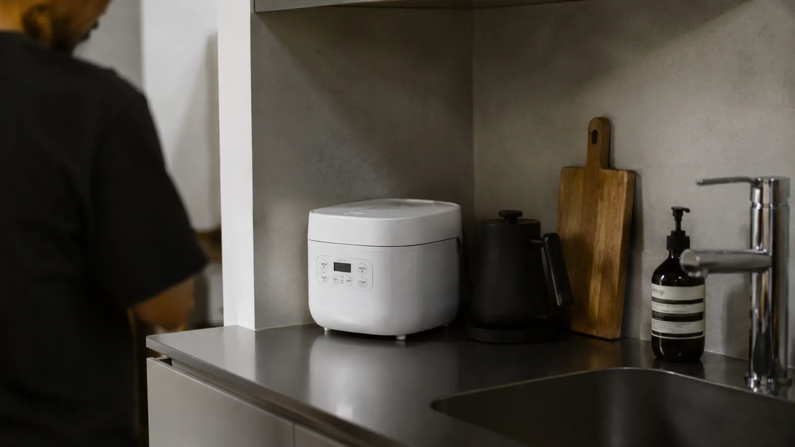 Person portioning cooked rice from a Dash Mini Rice Cooker into a meal-prep container in a small apartment kitchen